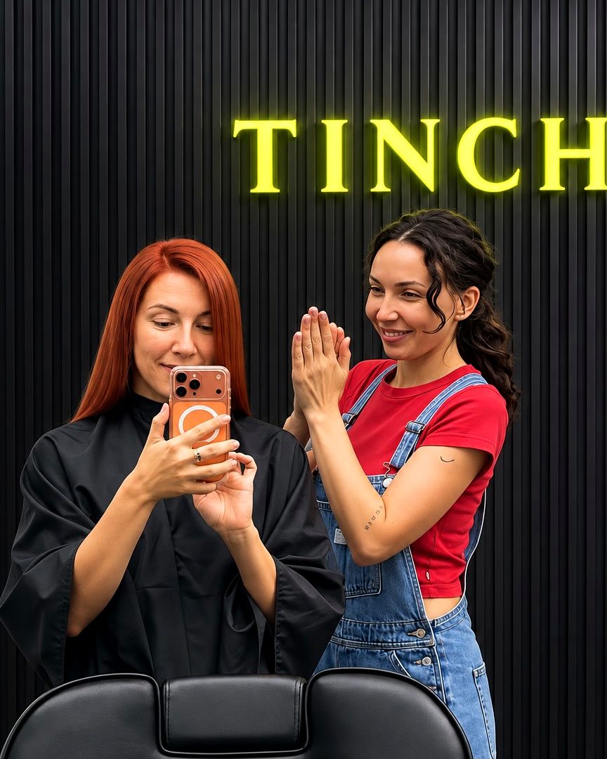 Tina applauding beside a happy red-haired client taking a mirror selfie of her fresh color, in front of the TinChik sign