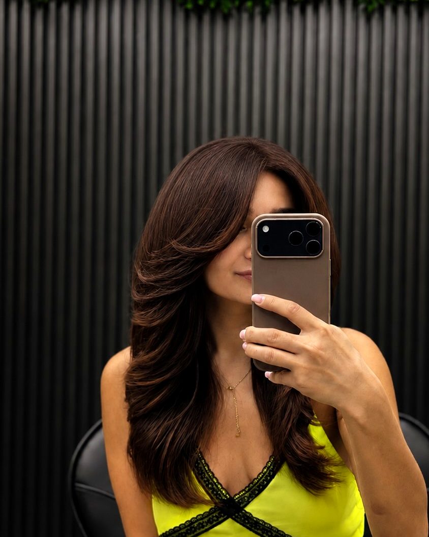 Client with long, glossy layered brunette hair taking a mirror selfie at TinChik